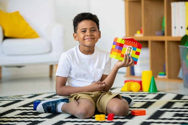 Mignon enfant d'âge préscolaire africain en tenue décontractée jouant sur le sol à la maternelle et la construction de la maison à partir de détails en plastique multicolores