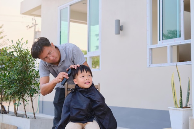 Photo mignon enfant asiatique souriant jeune garçon se faisant couper les cheveux dans la cour de la maison le père fait une coupe de cheveux pour son fils pendant le verrouillage coupe de cheveux à domicile pendant l'isolement en quarantaine pendant la crise sanitaire de covid19