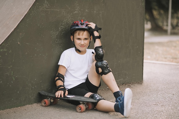 Mignon enfant garçon enfant dans un casque assis dans une zone spéciale du skatepark et tenant une planche à roulettes Concept d'activité sportive d'été Enfance heureuse