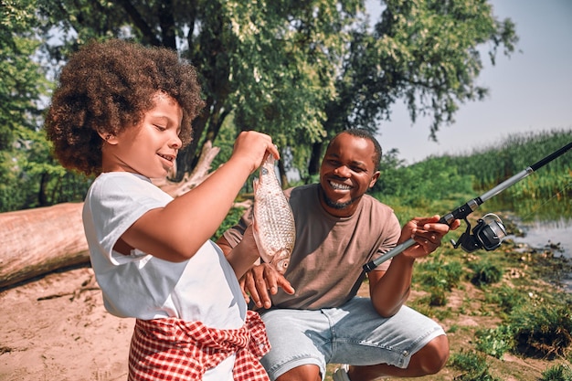 Mignon garçon afro-américain bouclé regardant le poisson dans ses mains pendant que son père tient une canne à pêche