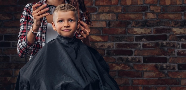 Mignon garçon d'âge préscolaire se coupe les cheveux. Le coiffeur pour enfants avec tondeuse coupe un petit garçon dans la chambre avec un intérieur loft.