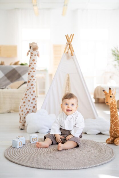 Un mignon garçon en bonne santé est assis sur un tapis dans le salon lumineux de la maison sur le fond d'un wigwam et de jouets en peluche jouant avec des jouets éducatifs en bois textiles de maison