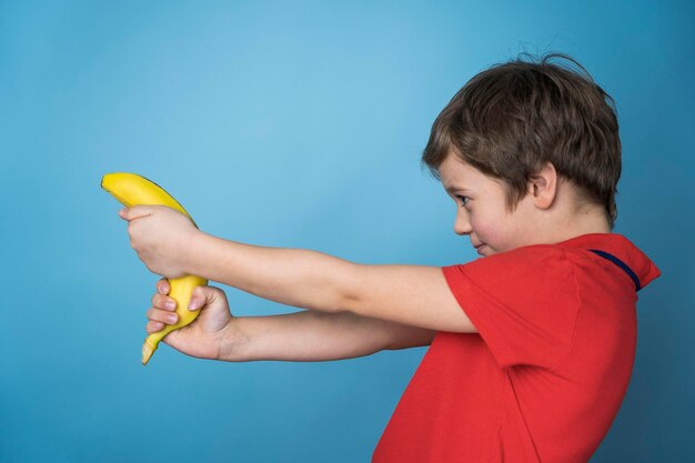 Un mignon garçon caucasien dans un T-shirt rouge tire agressivement et joyeusement une banane comme un pistolet.