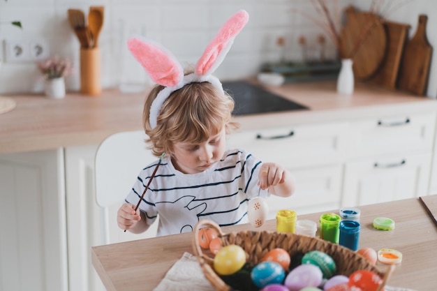 Mignon garçon de deux ans dans des oreilles de lapin peindre des oeufs de Pâques avec des peintures multicolores assis à la table de la cuisine