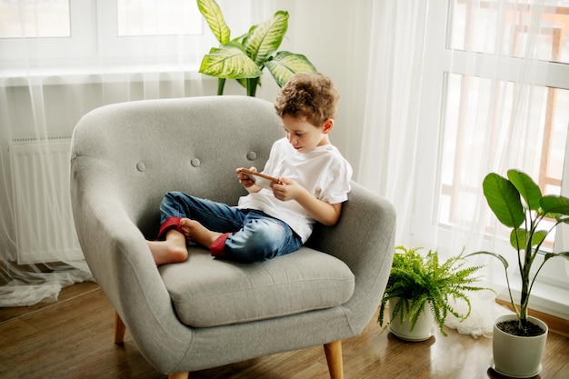 Un mignon garçon est assis dans un fauteuil et joue à un jeu sur son téléphone ou regarde des vidéos éducatives