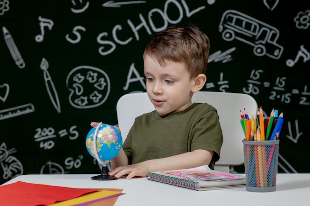 Mignon garçon intelligent est assis à un bureau avec globe à la main sur fond avec tableau noir. Prêt pour l'école. Retour à l'école