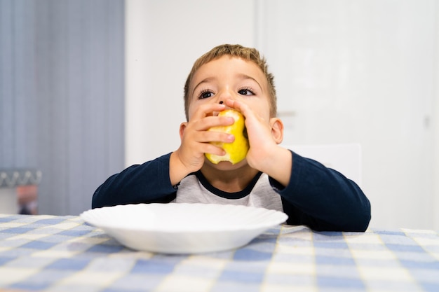 Un mignon garçon mangeant une pomme alors qu'il est assis à la table à la maison.