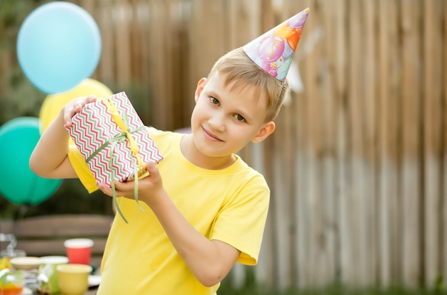 Mignon garçon de neuf ans drôle célébrant son anniversaire avec sa famille ou ses amis dans une cour Fête d'anniversaire Enfant portant un chapeau de fête et tenant une boîte-cadeau