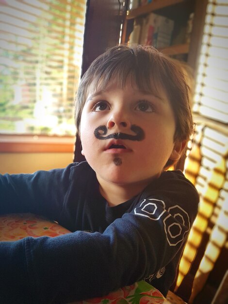 Photo un mignon garçon avec la peinture sur le visage qui détourne son regard alors qu'il est assis à la table à la maison.