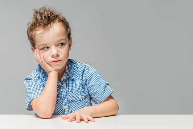 Un mignon garçon réfléchi en chemise bleue à la table sur un fond gris isolé