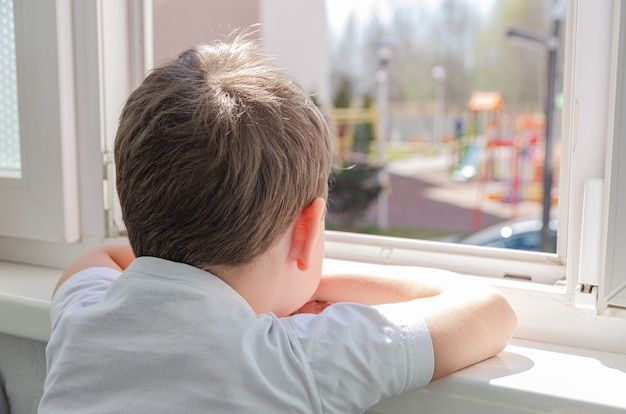 Mignon garçon triste de quatre ans est assis sur le balcon et regarde par la fenêtre de l'aire de jeux. Auto-isolement (quarantaine)