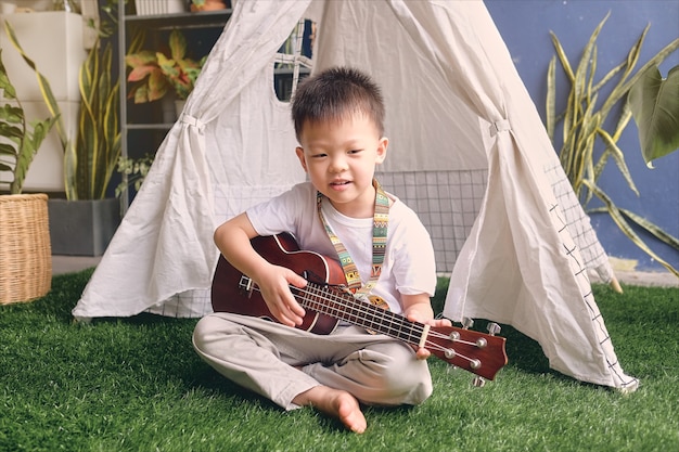 Mignon heureux souriant petit garçon asiatique de maternelle enfant s'amusant à jouer de la guitare hawaïenne ou du ukulélé