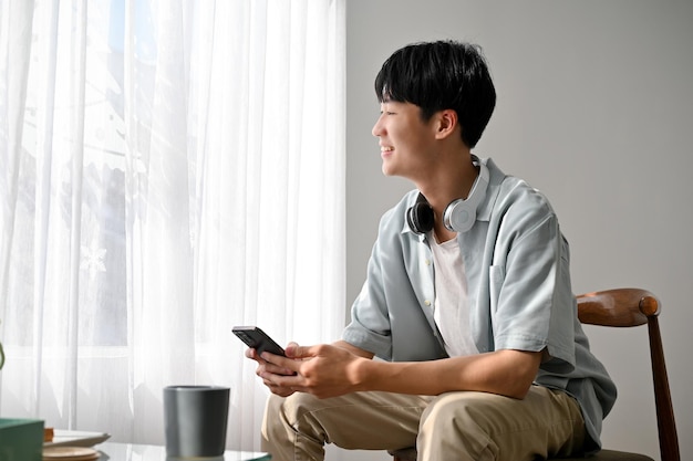 Mignon jeune homme asiatique tenant un smartphone assis sur une chaise en regardant par la fenêtre