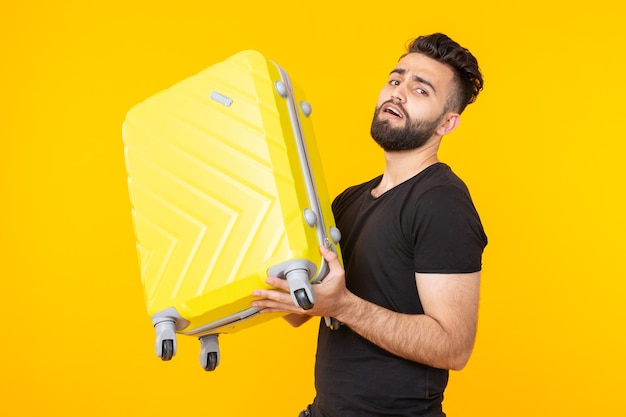 Mignon jeune homme barbu hipster tenant une lourde valise jaune sur fond jaune.