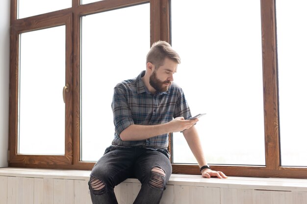 Mignon jeune homme hipster avec des vêtements élégants est assis sur les réseaux sociaux à l'aide d'un smartphone et