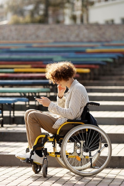 Mignon mec handicapé aux cheveux bouclés en fauteuil roulant regarde le téléphone Un jeune homme passe son temps libre à l'extérieur