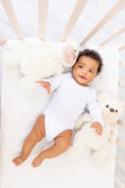 Mignon petit bébé afro-américain dans un lit de couchage blanc avec des jouets d'ours.