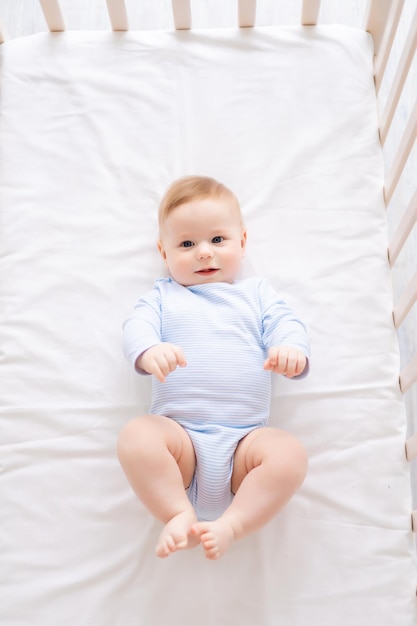 Un mignon petit bébé en bonne santé est allongé sur le dos dans un berceau sur une literie blanche à la maison dans un body bleu L'enfant heureux regarde la caméra sourit