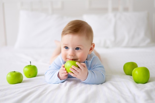 Un mignon petit bébé en bonne santé est allongé sur un lit sur une literie blanche à la maison dans un body bleu Le bébé tient dans ses mains et goûte les pommes vertes le premier aliment complémentaire des aliments sains