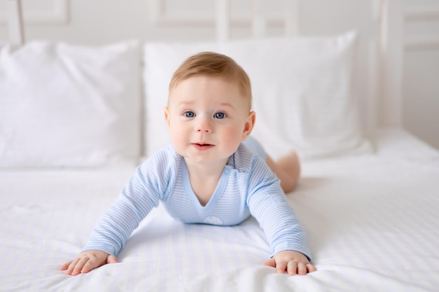 Un mignon petit bébé en bonne santé est allongé sur un lit sur une literie blanche à la maison dans un body bleu L'enfant regarde la caméra sourit