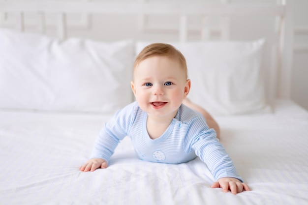 Un mignon petit bébé en bonne santé est allongé sur un lit sur une literie blanche à la maison dans un body bleu L'enfant regarde la caméra sourit