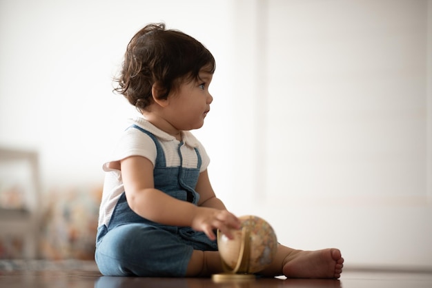 Mignon petit bébé garçon tout-petit assis sur un plancher en bois jouant avec un globe terrestre tout en regardant ailleurs et en pensant à un jouet cassé