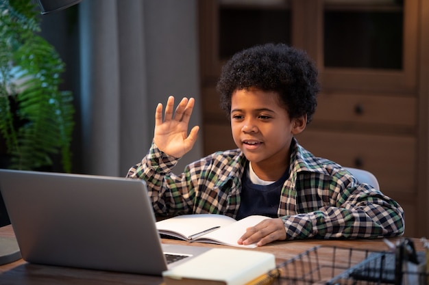 Mignon petit écolier africain en tenue décontractée agitant la main alors qu'il était assis près d'une table devant un ordinateur portable, regardant l'affichage et saluant son professeur