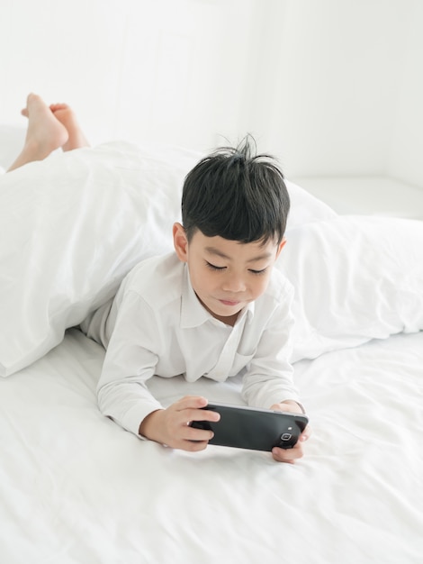 Mignon petit enfant asiatique concentré sur un smartphone en position couchée sur le lit