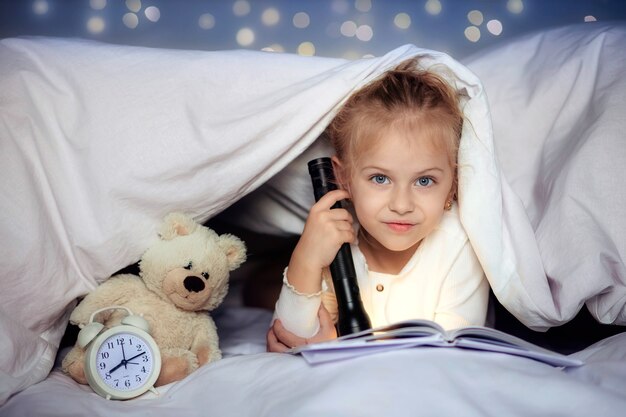 Mignon petit enfant lisant un livre avec une lampe de poche sous la couverture