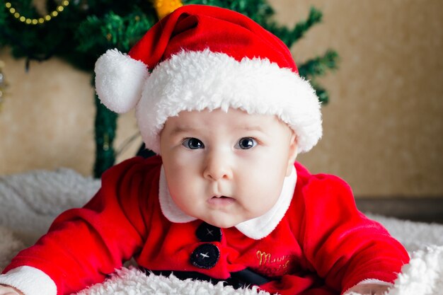 Mignon petit enfant de Noël vêtu d'un costume de père Noël et regardant le spectateur, arbre de Noël au mur