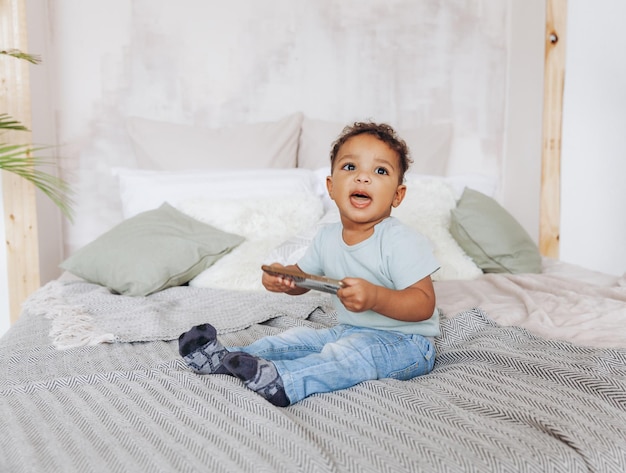 Mignon petit garçon afro-américain avec smartphone à la maison sur le lit heureux enfant regardant des dessins animés au téléphone