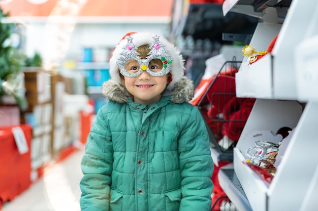Mignon petit garçon d'âge préscolaire portant un chapeau de Noël et un masque choisissant la décoration dans un supermarché