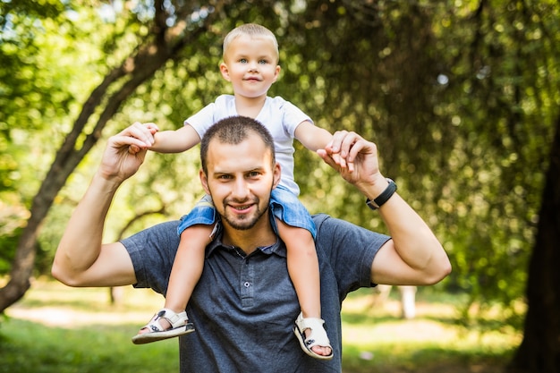 Photo mignon petit garçon assis pickaback de son beau jeune papa se regardent et souriant tout en se reposant dans le parc