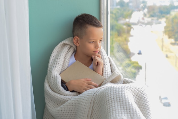 Mignon petit garçon assis près de la fenêtre et lisant un livre dans la chambre à la maison Belle nature d'automne