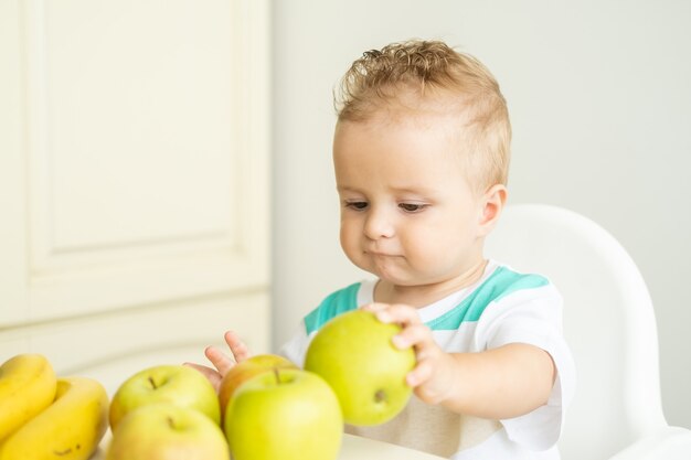 Mignon petit garçon assis à la table dans une chaise enfant mangeant une pomme sur une cuisine blanche.