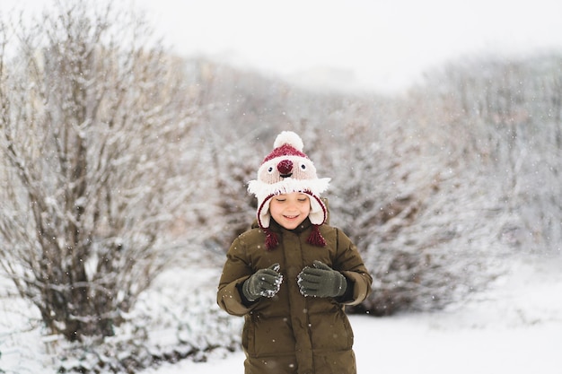 Mignon petit garçon au chapeau d'hiver drôle se promène pendant une chute de neige en plein air activités d'hiver pour les enfants