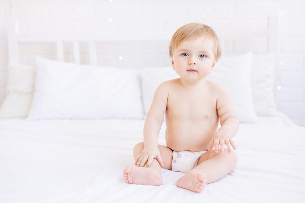 Mignon petit garçon blond assis sur un lit blanc à la maison en couches dans une pièce lumineuse