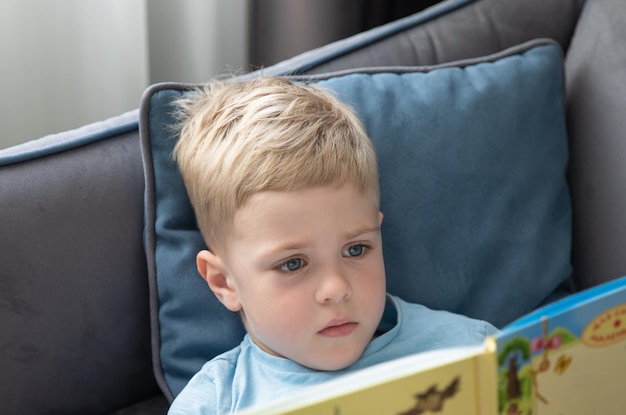 Mignon petit garçon blond lisant un livre allongé sur le canapé