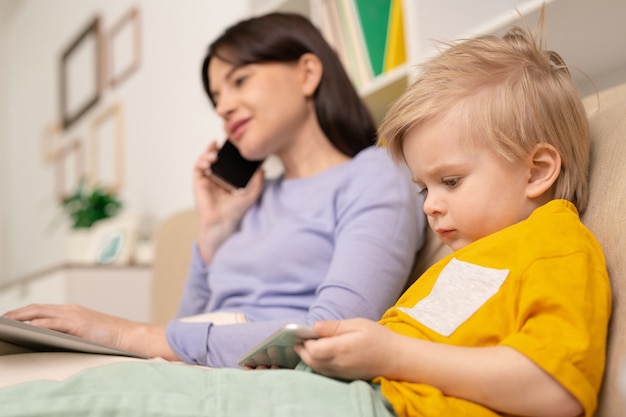 Mignon Petit Garçon Blond Avec Smartphone Regardant Un Dessin Animé En Ligne Pendant Que Sa Mère Parle Au Téléphone Et Travaille à Distance En Quarantaine