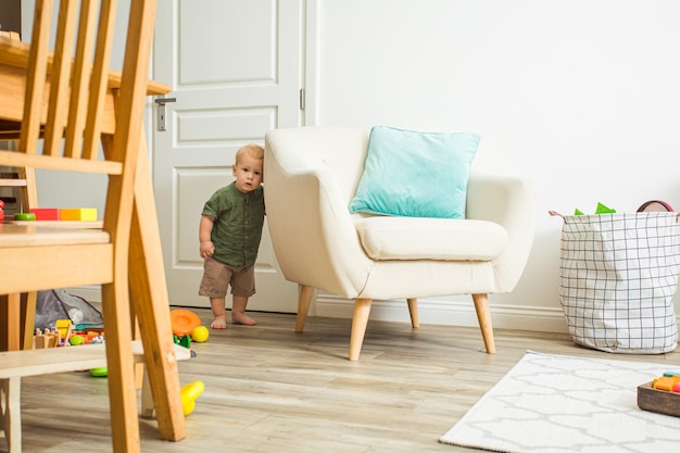 Mignon petit garçon blond timide se cachant près d'un grand fauteuil blanc honteux de venir rencontrer des invités L'enfant apprend à communiquer avec de nouvelles personnes dans sa vie Premier jour avec nounou