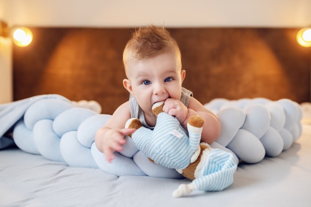 Mignon petit garçon caucasien joufflu couché sur le ventre sur le lit dans la chambre et mâcher l'ours en peluche. La première dent est presque là.