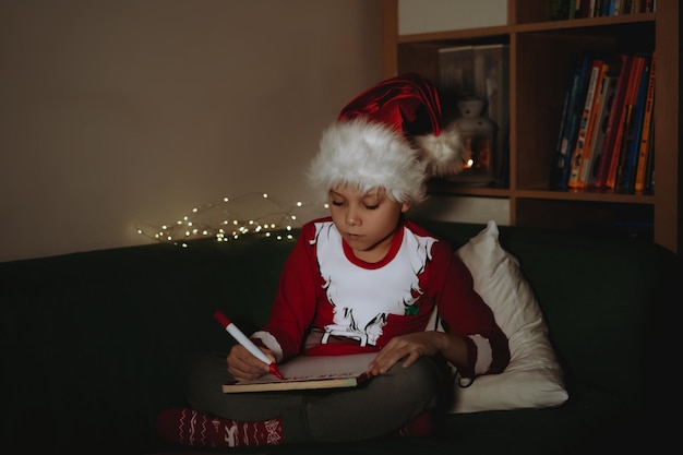 Mignon petit garçon caucasien en santa tee shirt et lettre de hatwriting au Père Noël assis sur un canapé. photo de haute qualité