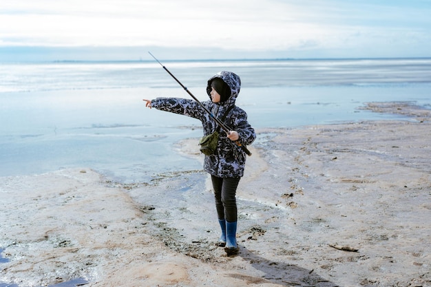 Mignon petit garçon caucasien tenant une canne à pêche regardant à distance au bord de la mer le jour du printemps