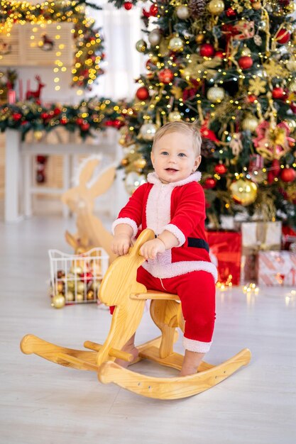 Un mignon petit garçon en costume de Père Noël chevauche un jouet à bascule sous un arbre de Noël festif avec des cadeaux dans le salon de la maison un enfant heureux fête Noël et le Nouvel An à la maison