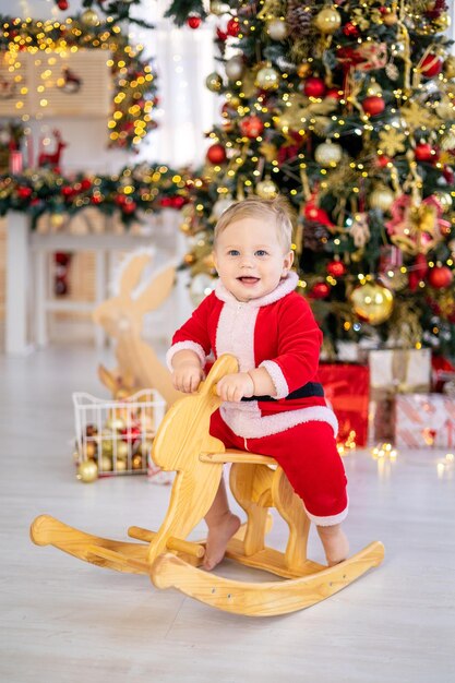 Un mignon petit garçon en costume de Père Noël chevauche un jouet à bascule sous un arbre de Noël festif avec des cadeaux dans le salon de la maison un enfant heureux fête Noël et le Nouvel An à la maison