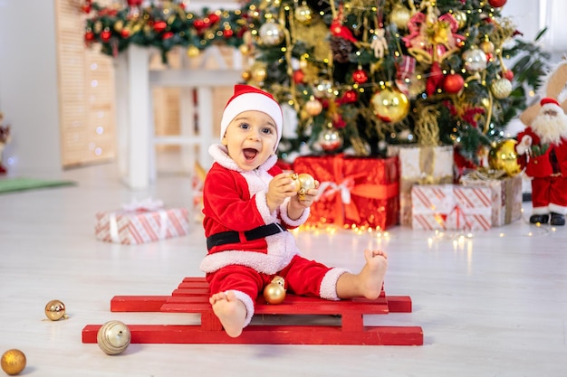 Un mignon petit garçon en costume de Père Noël est assis sur un traîneau sous un arbre de Noël festif avec des cadeaux dans le salon de la maison un enfant heureux célèbre Noël et le Nouvel An à la maison
