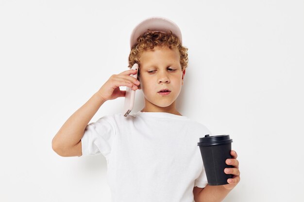 Mignon petit garçon dans une casquette de t-shirt blanc avec un téléphone dans un verre avec une boisson fond clair inchangé