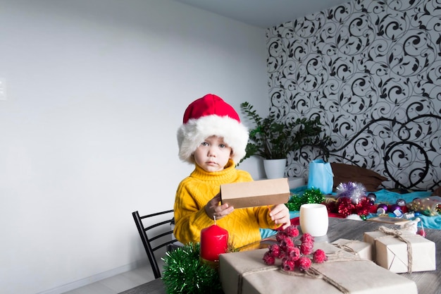 Un mignon petit garçon dans un chandail jaune et un chapeau de Père Noël rouge sur une table en bois avec des cadeaux emballés dans du papier kraft