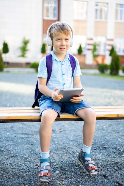 Mignon petit garçon dans des écouteurs et des tablettes sans fil assis sur un banc dans la cour de l'école d'apprentissage à distance