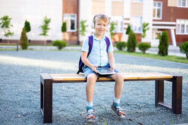 Mignon petit garçon dans des écouteurs et des tablettes sans fil assis sur un banc dans la cour de l'école d'apprentissage à distance